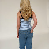 En person med langt, blondt, bølget hår står med ryggen til, iført en stribet tanktop og jeans, på en neutral, lys baggrund.