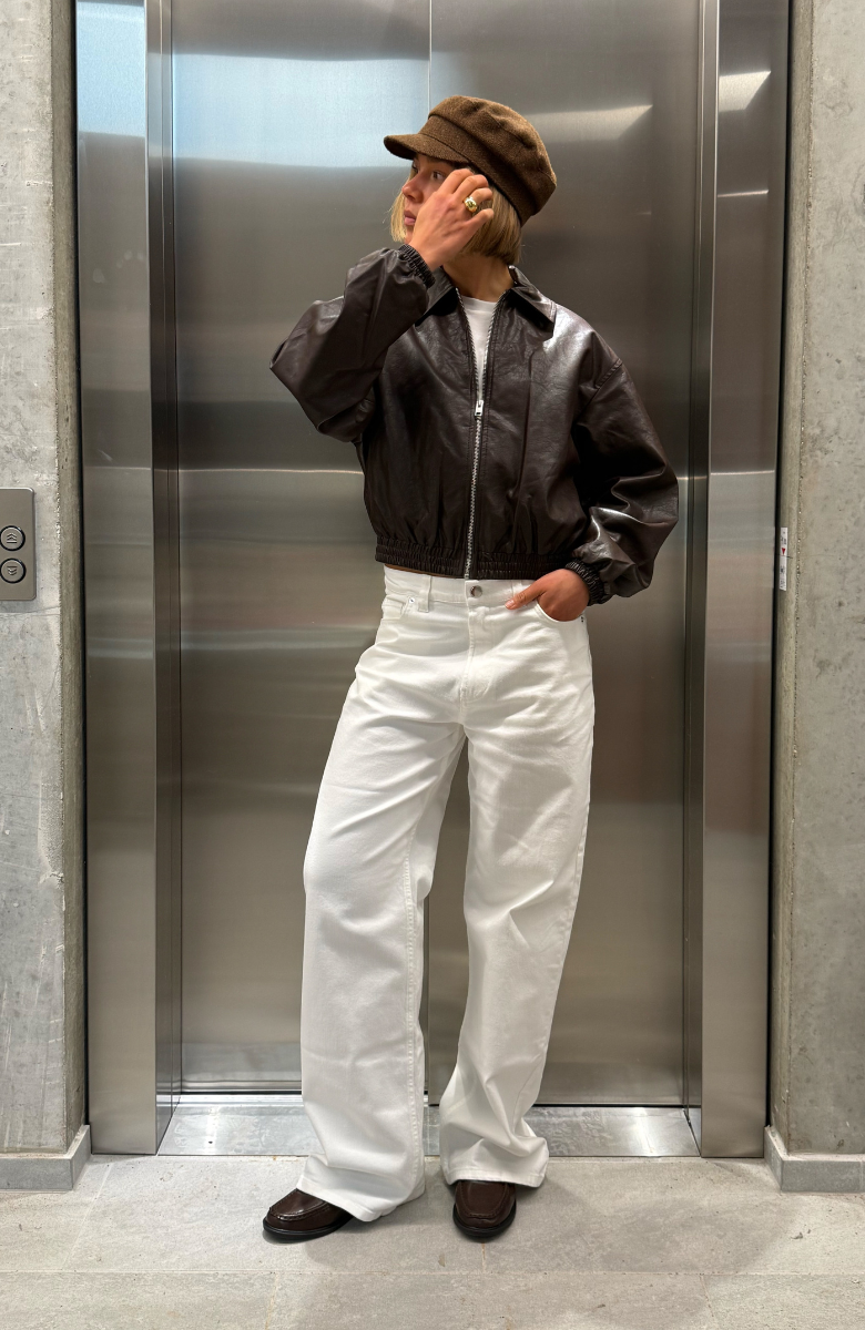 Stemningsbillede af pige ved elevatordør. Pigen bærer hvide NANNA jeans og brun læderjakke. Fra BySofieSønderby