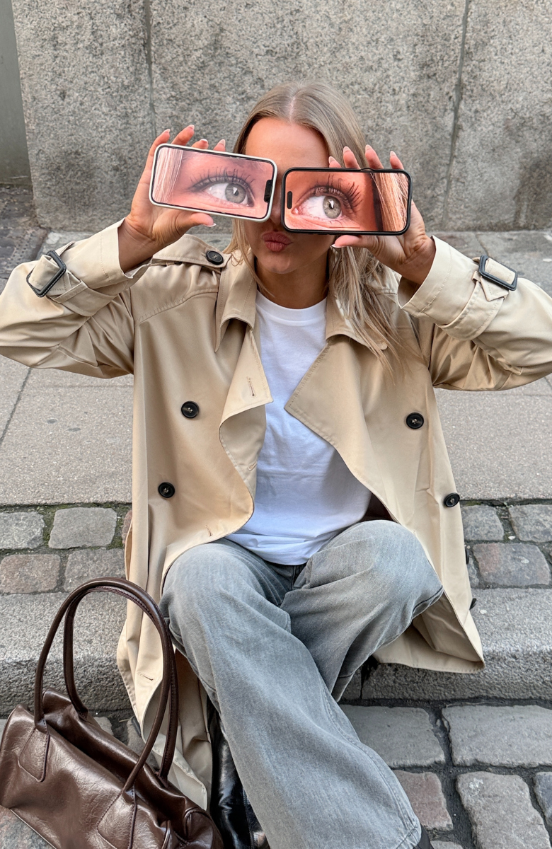 Stemningsbillede af pige i beige trenchcoat fra BySofieSødnerby. Hun sidder med to mobiler som øjne.