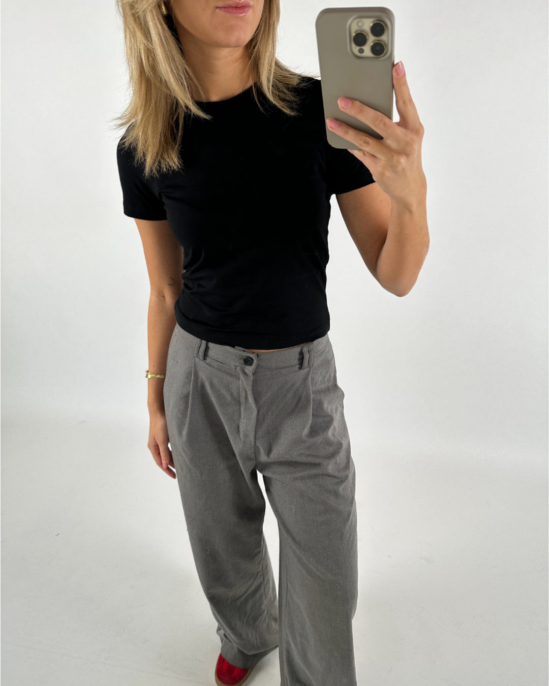 Person tager et spejlbillede iført sort T-shirt, grå bukser og røde sko i et minimalistisk rum. Personen holder en smartphone med et lysegråt cover.