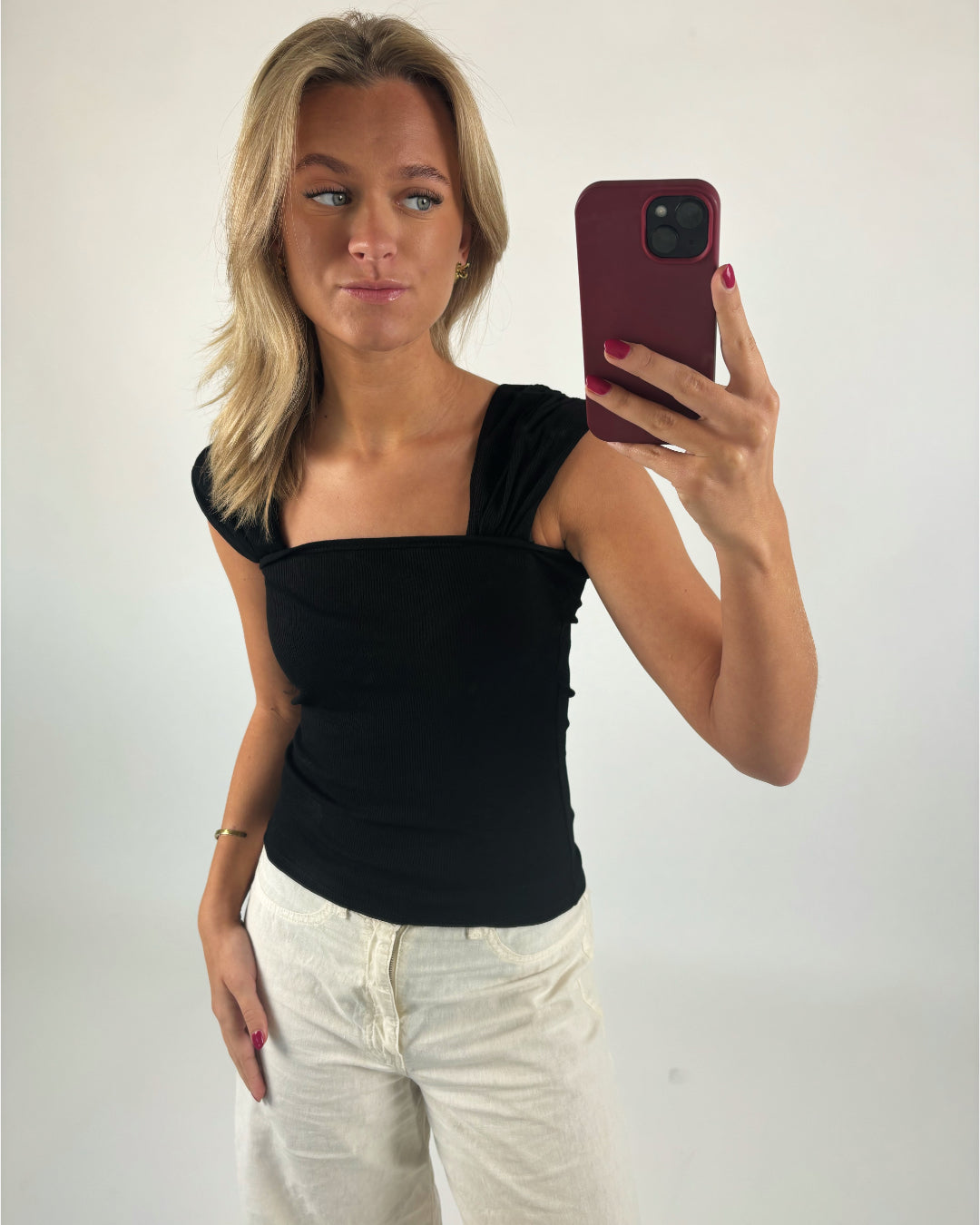 En person tager en selfie med en rød telefon i hånden, iført en sort top og hvide bukser, i et neutralt belyst rum.