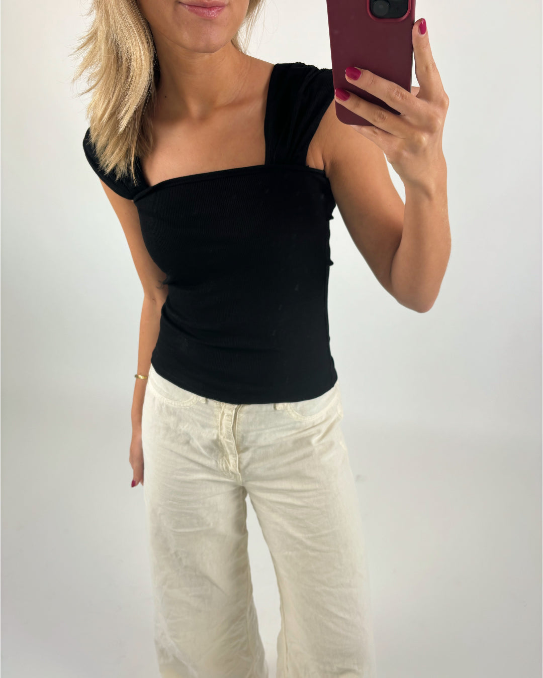 Person tager selfie; iført sort top og hvide bukser; indendørs med hvid baggrund.
