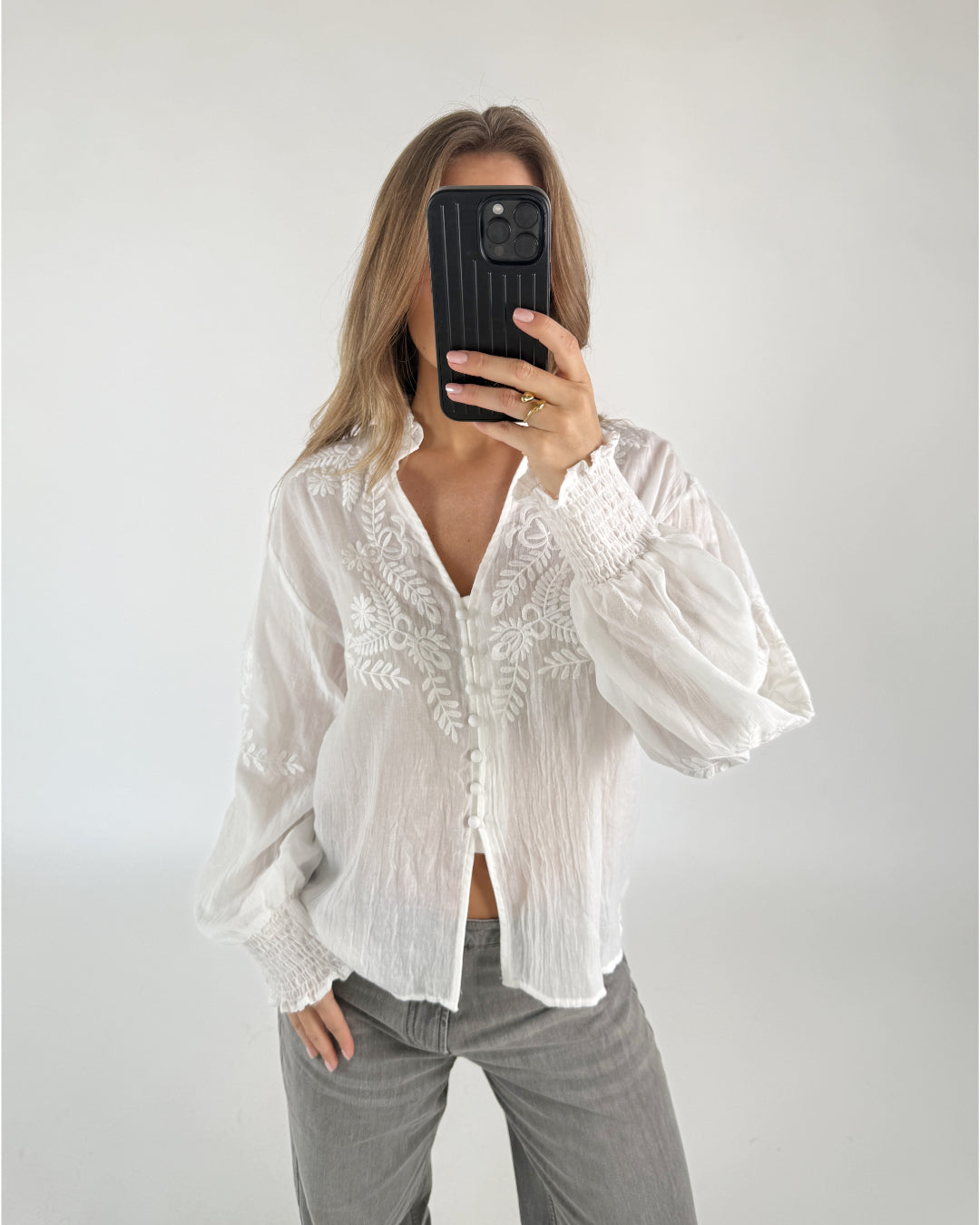 En person tager et spejlfoto, iført en hvid bluse med broderi og grå bukser, i et lyst rum. Personen holder en mobiltelefon op foran ansigtet.
