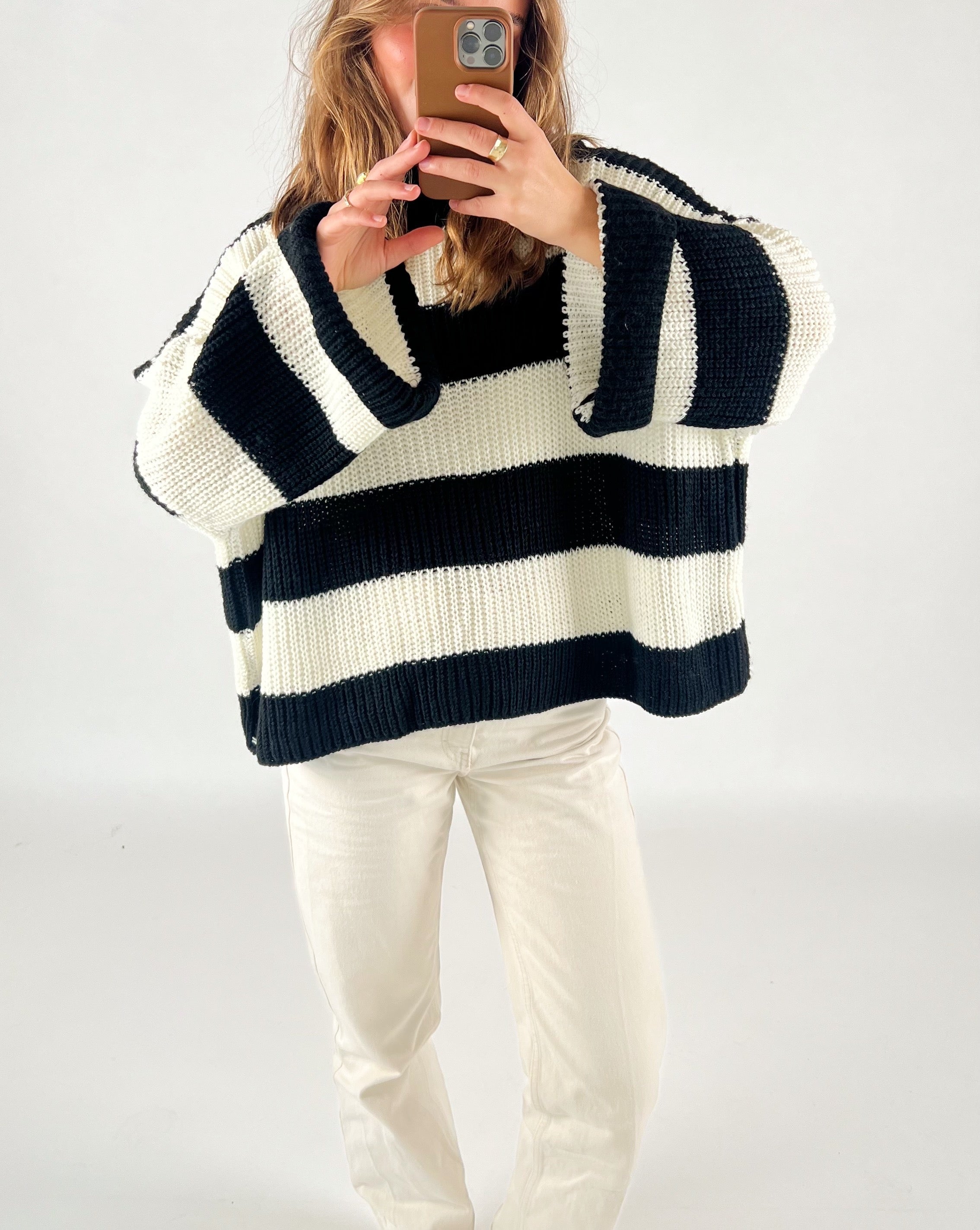 En person iført en sort-hvidstribet sweater tager et selfie i et spejl. Personen står i et lyst rum og bærer hvide bukser.