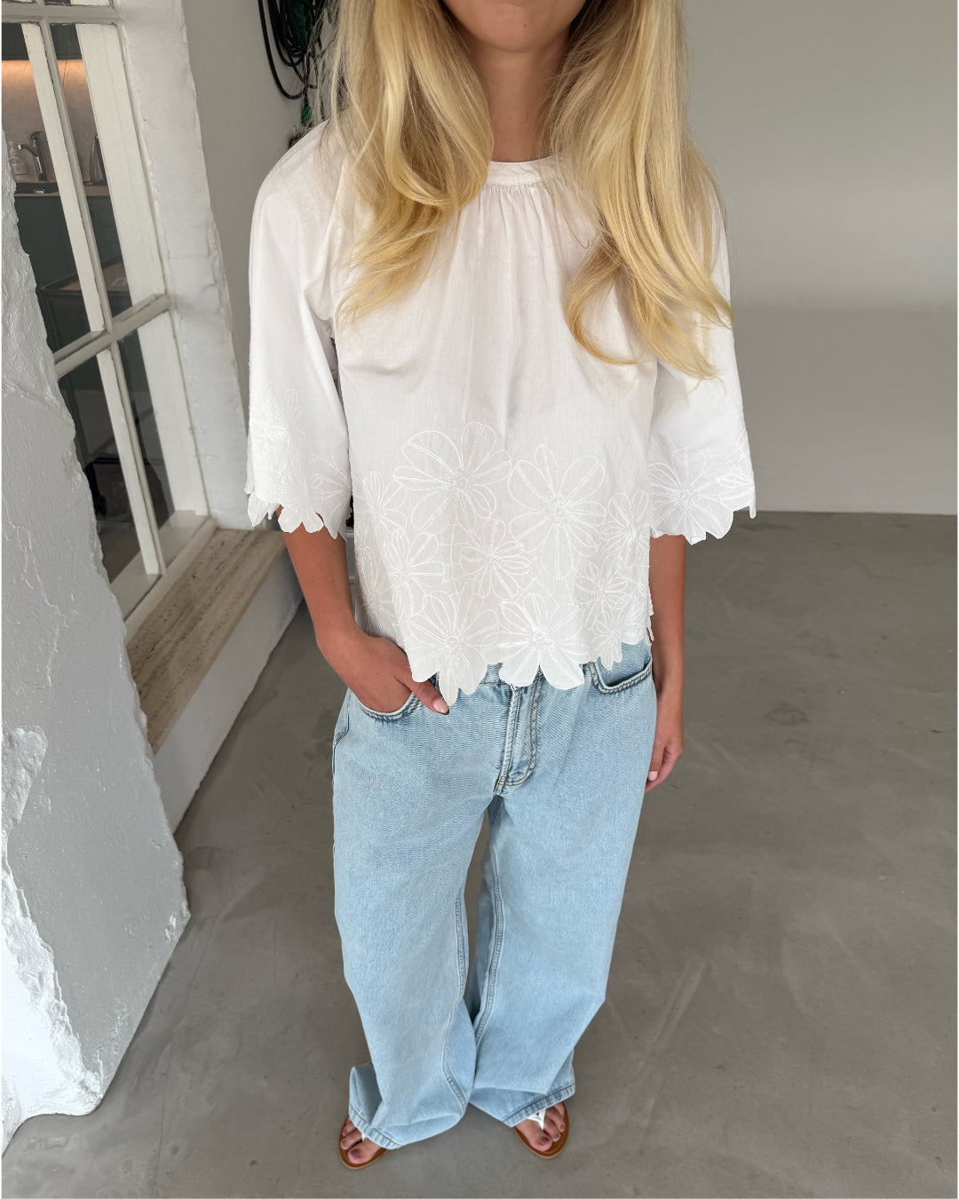 Modelfoto af pige som bærer hvid KAMMA bluse med blomstret sy-mønster, samt lyseblå jeans. Alt er fra BySofieSønderby