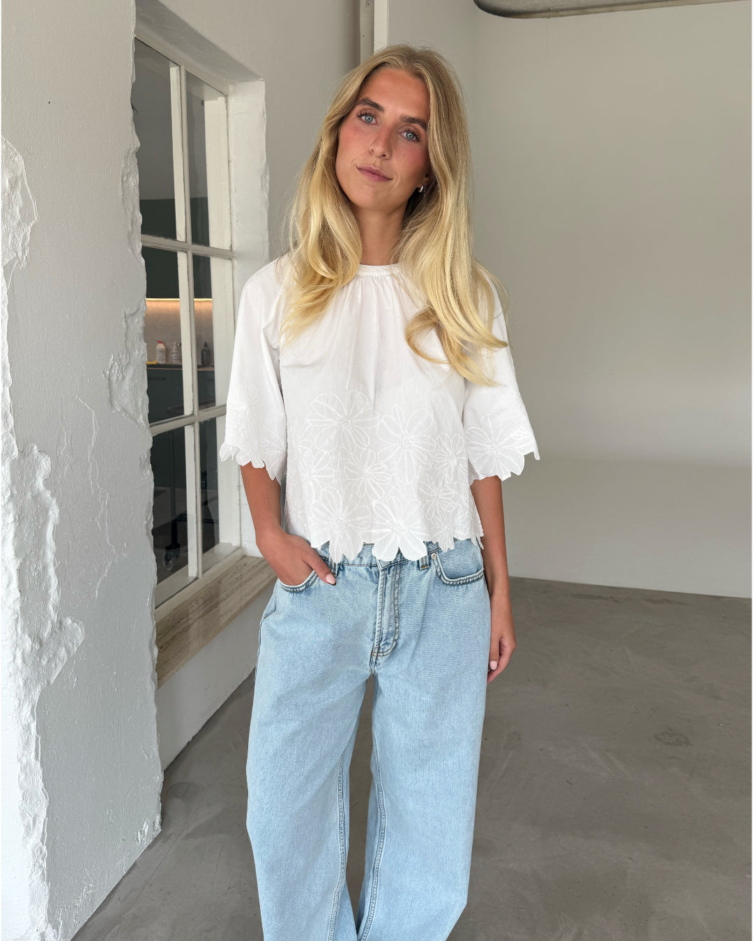 Modelfoto af pige som bærer hvid KAMMA bluse med blomstret sy-mønster, samt lyseblå jeans. Alt er fra BySofieSønderby