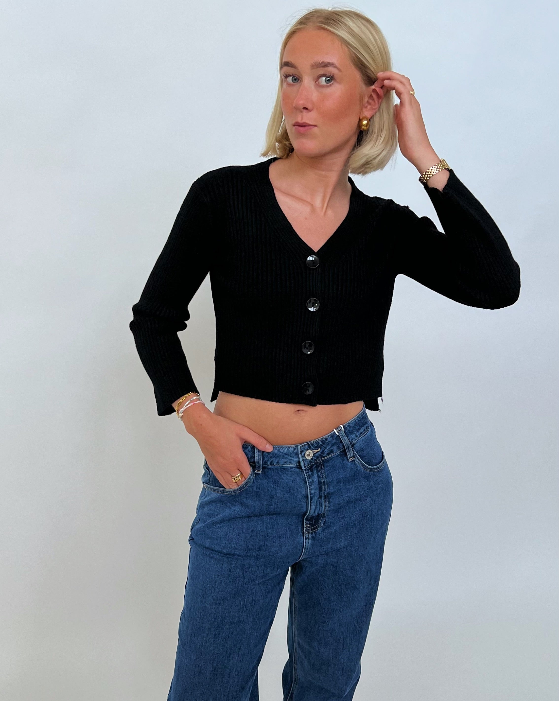 En person i sort cardigan og jeans rører ved sit hår, mens de står mod en neutral baggrund.