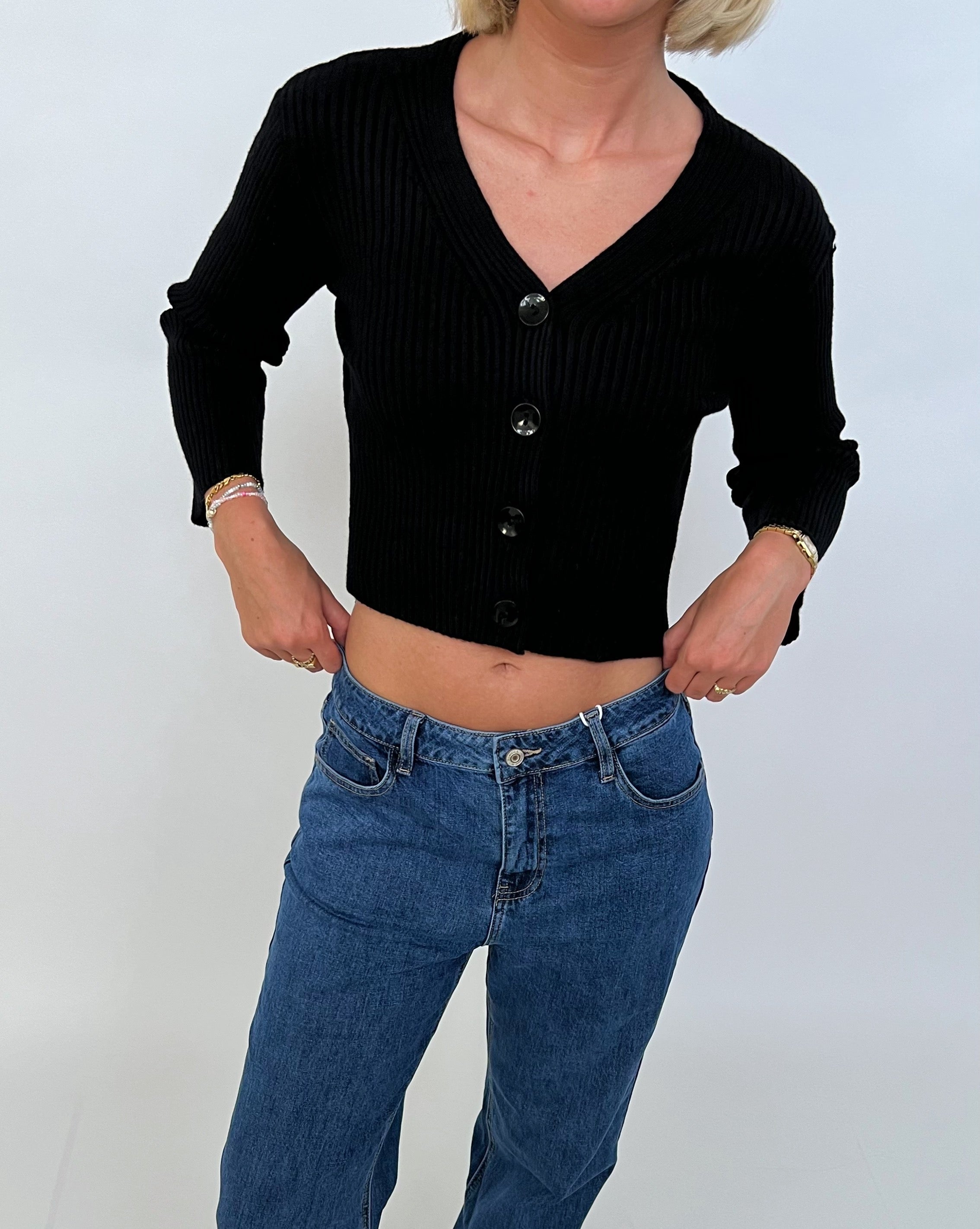 Kvinde bærer sort cardigan med knapper og blå jeans, står i afslappet pose med hænderne i lommerne, mod en simpel hvid baggrund.