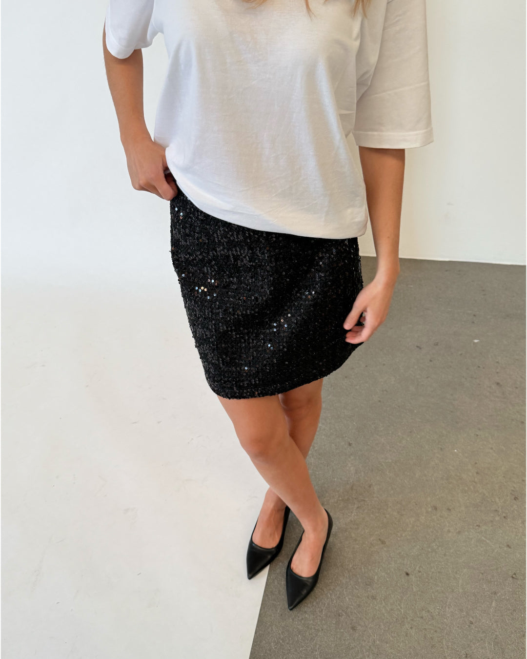 Modelfoto af pige i sort KENDALL nederdel med pailetter, samt hvid basic t-shirt og spidse ballerinaer. Alt er fra BySofieSønderby.