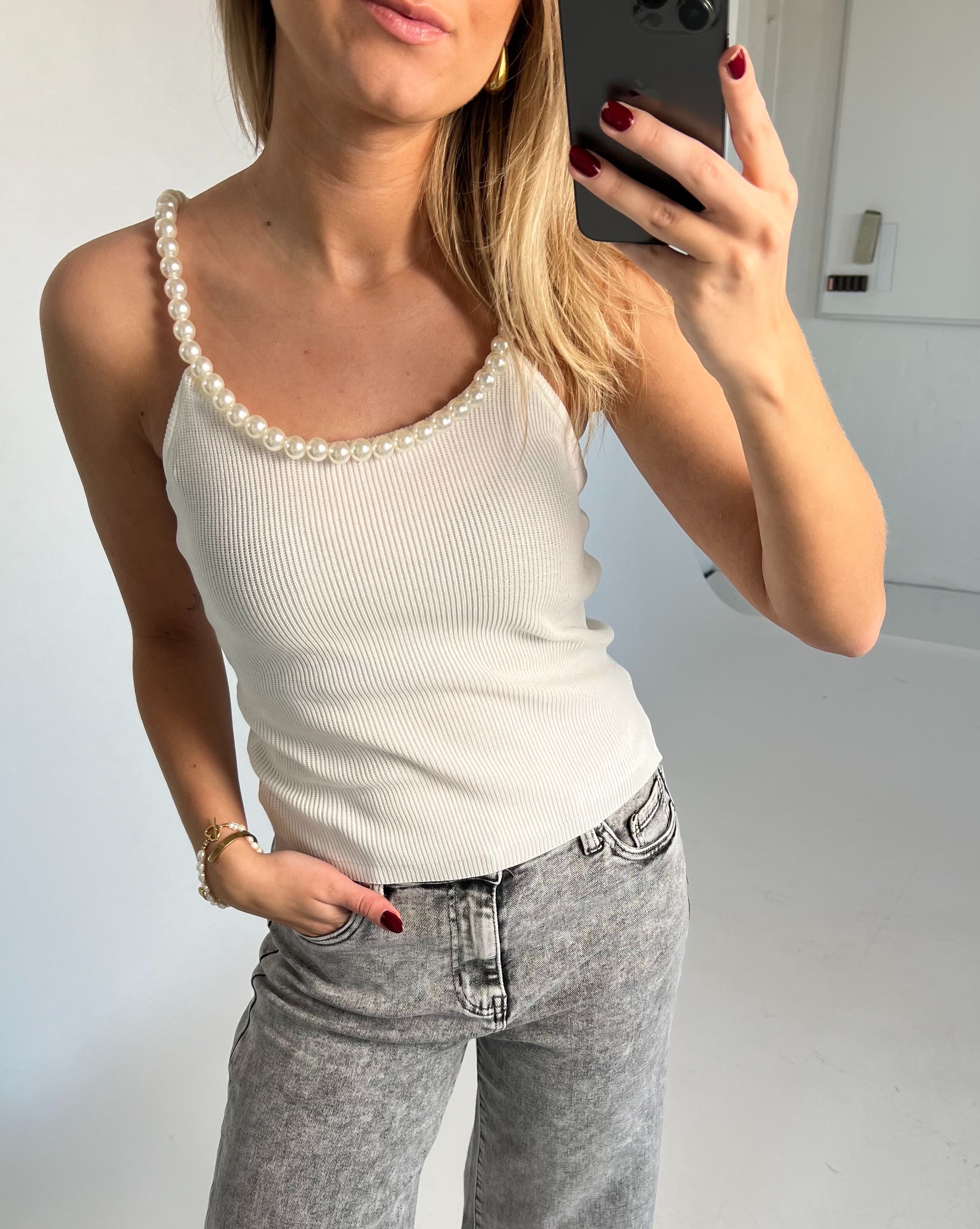 En person holder en telefon, tager et billede af sig selv iført en hvid top med perlekant og grå jeans. Baggrunden er enkel og lys.