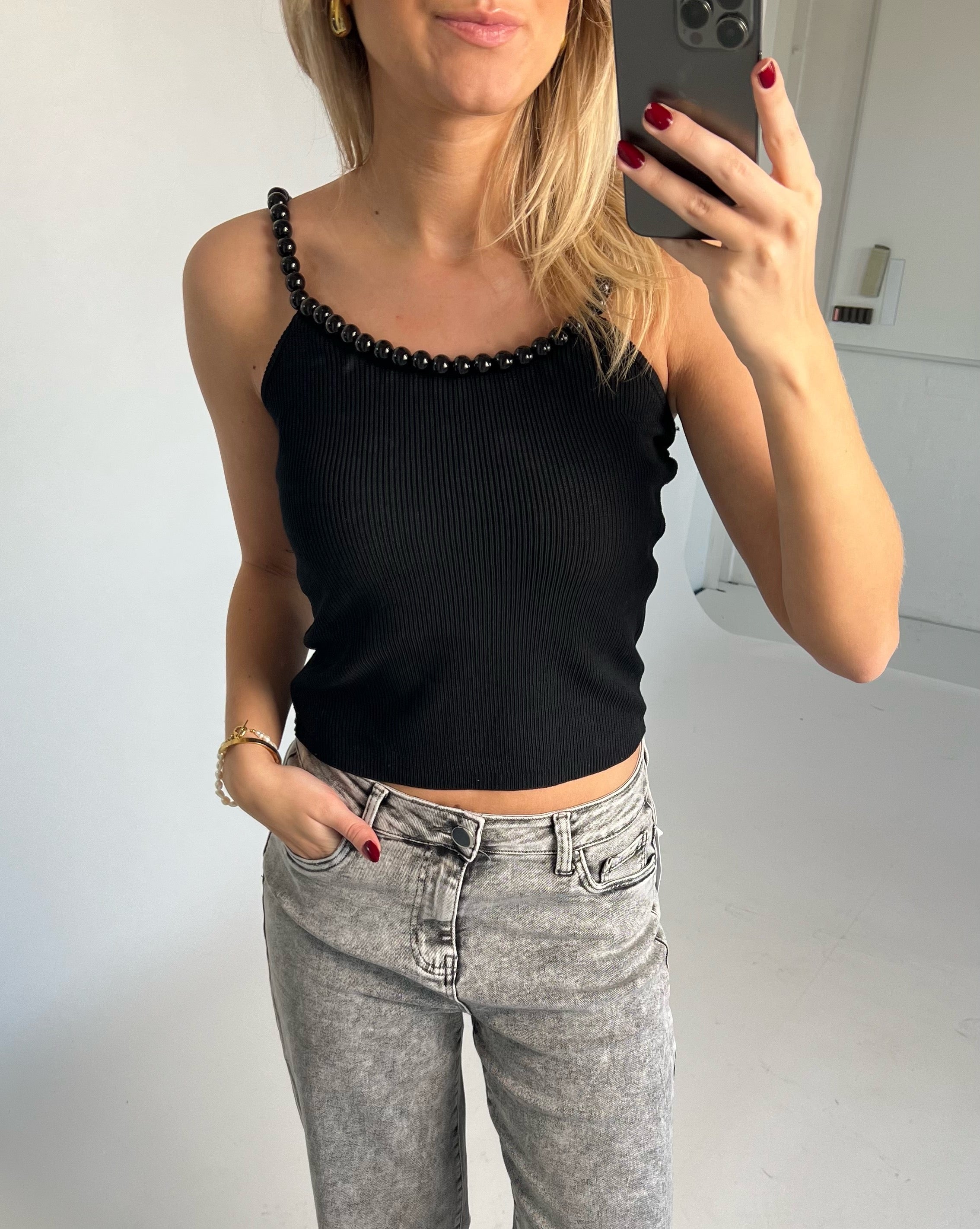 Kvinde iført sort top med perlepynt tager selfie. Hun har lyse jeans og rød neglelak. Baggrund er et lyst indendørs rum.