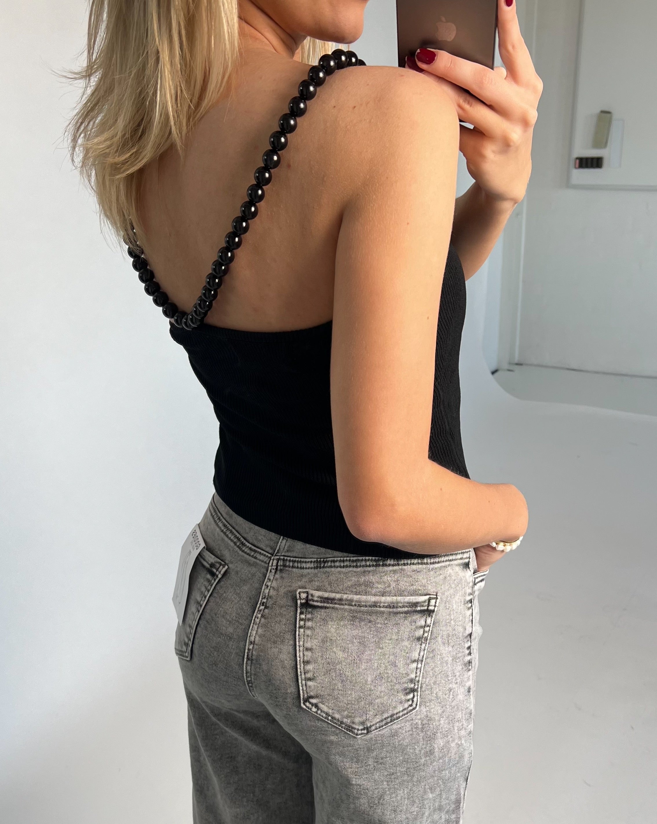 En kvinde iført en sort top med perlestropper og grå jeans tager et spejlbillede i et lyst rum.