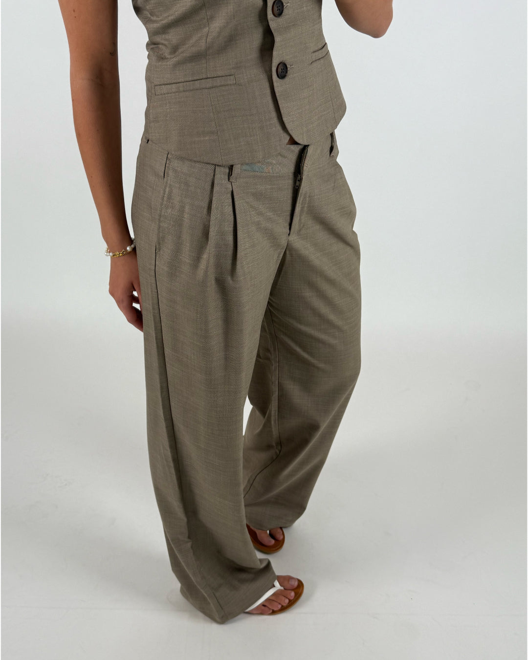 En person iført en beige vest og bukser står med hænderne afslappet ved siden, mod en neutral baggrund. Personen bærer sandaler og et armbånd.