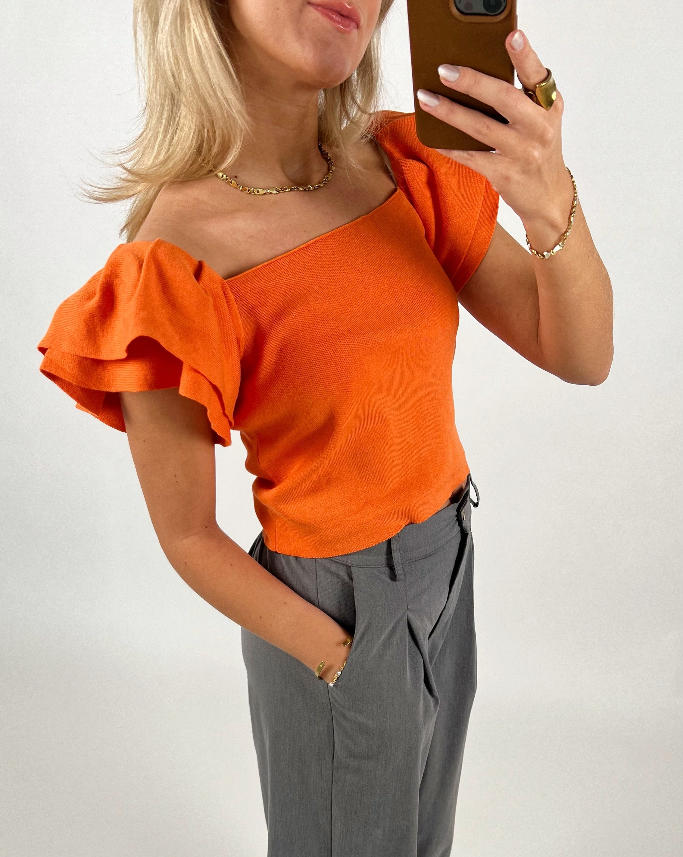 En person iført en orange top med puffærmer tager et spejlbillede. Personen bærer grå bukser og smykker. Baggrunden er simpel og hvid.