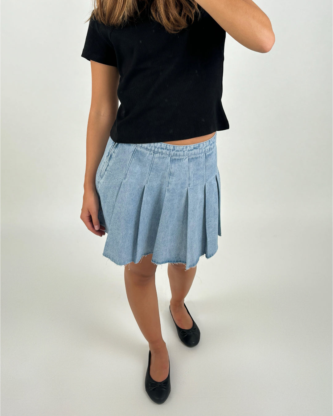 En person iført sort t-shirt og lys denimnederdel står i et neutralt, hvidt rum. Personen har sorte sko på og har en hånd ved siden af kroppen.
