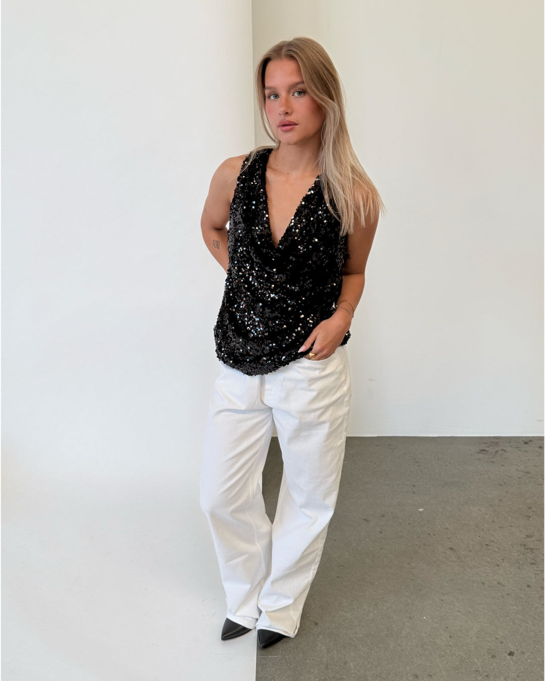 Modelfoto af pige i sort LINA top med pailetter, samt hvide NANNA jeans. Alt er fra BySofieSønderby.