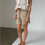 Person iført beige shorts og hvid bluse med flæser, stående på krydsfelt mellem gråt og hvidt underlag, indendørs fotograferet.