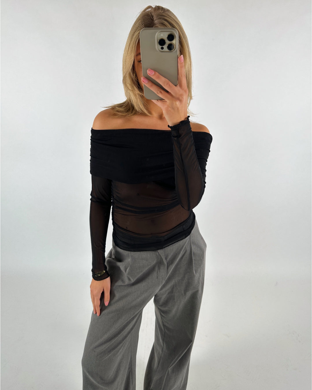 En person i en sort, off-shoulder top tager en selfie med en smartphone. Personen har grå bukser på, og baggrunden er en neutral, hvid væg.