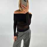 En person i en sort, off-shoulder top tager en selfie med en smartphone. Personen har grå bukser på, og baggrunden er en neutral, hvid væg.