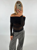 En person i en sort, off-shoulder top tager en selfie med en smartphone. Personen har grå bukser på, og baggrunden er en neutral, hvid væg.