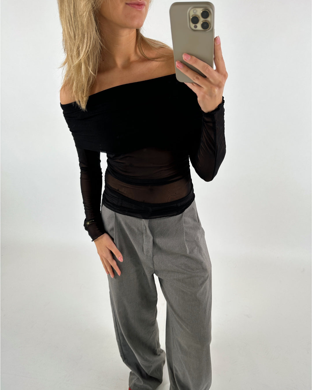 En person iført en sort off-shoulder trøje og grå bukser tager et spejlbillede med en smartphone i et minimalistisk rum.
