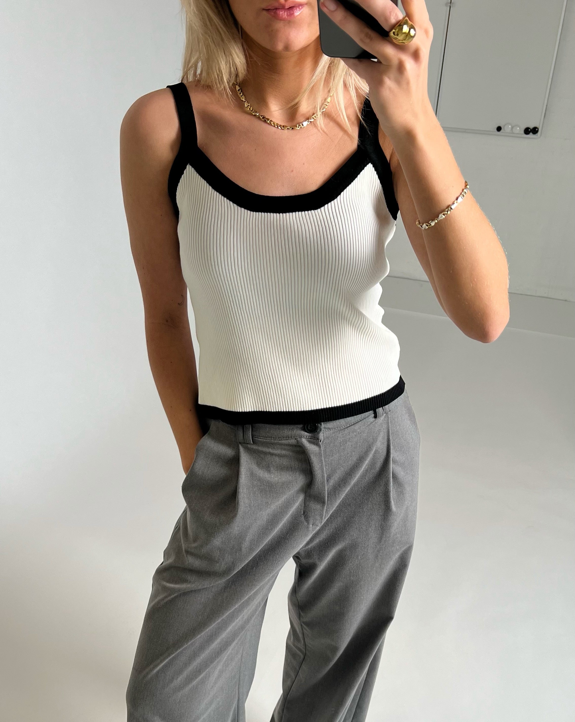 En person iført en hvid top med sorte kanter og grå bukser står foran et spejl og tager et billede med en smartphone i en minimalistisk, lys indendørs setting.