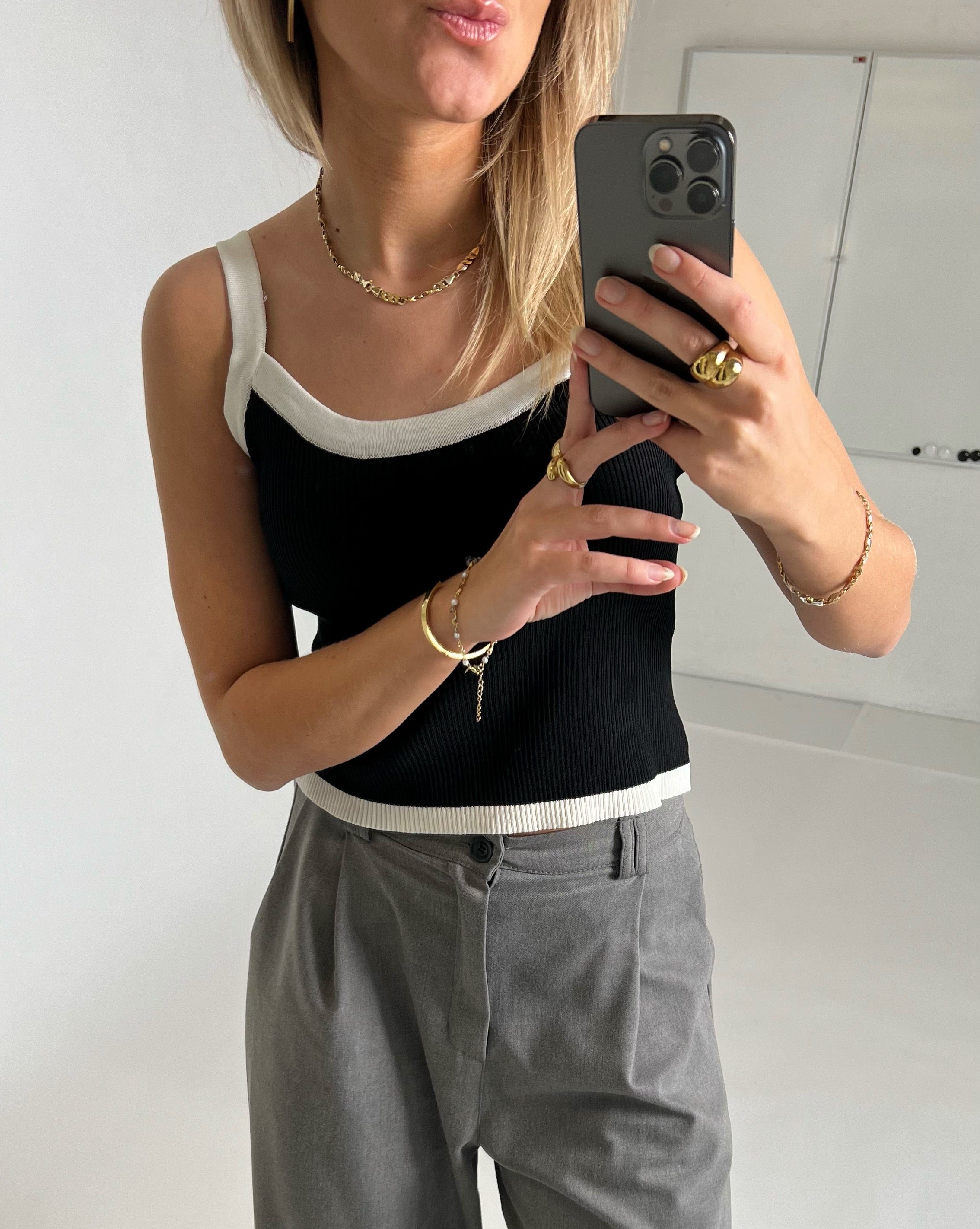 En person tager en selfie i et spejl, iført en sort top og grå bukser, iført guldsmykker og holder en sort smartphone, i et lyst rum uden tekst.