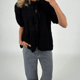 En person tager et spejlbillede, iført sort, mønsterstrikket cardigan og grå jeans. Personen står i et lyst rum og holder en smartphone.