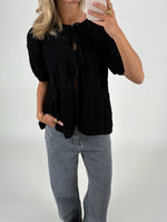 En person tager et spejlbillede, iført sort, mønsterstrikket cardigan og grå jeans. Personen står i et lyst rum og holder en smartphone.