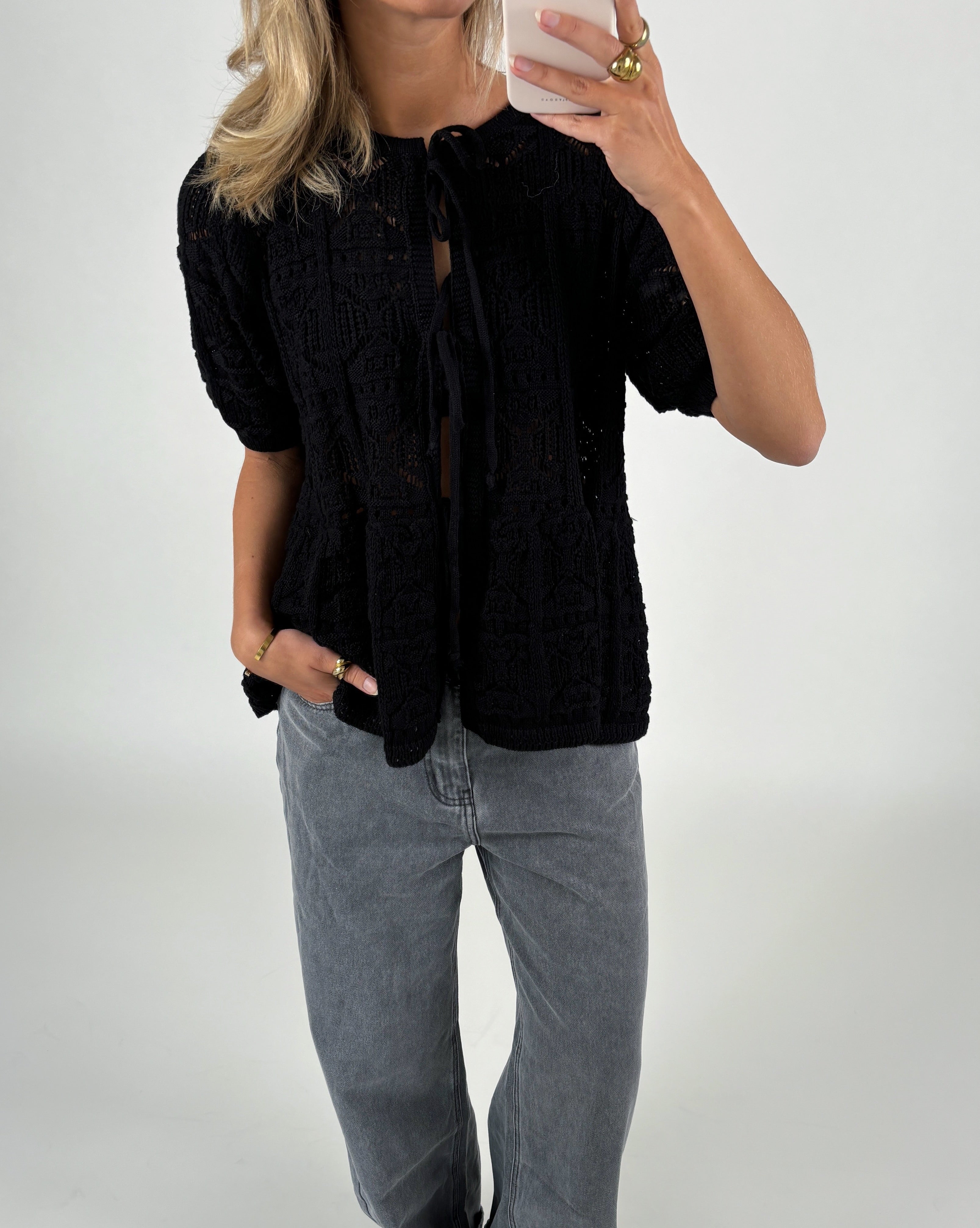 En person tager et spejlbillede, iført sort, mønsterstrikket cardigan og grå jeans. Personen står i et lyst rum og holder en smartphone.