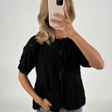 Person tager et spejlbillede iført sort striktrøje og grå jeans, med en smartphone foran ansigtet. Baggrunden er ensfarvet lys.