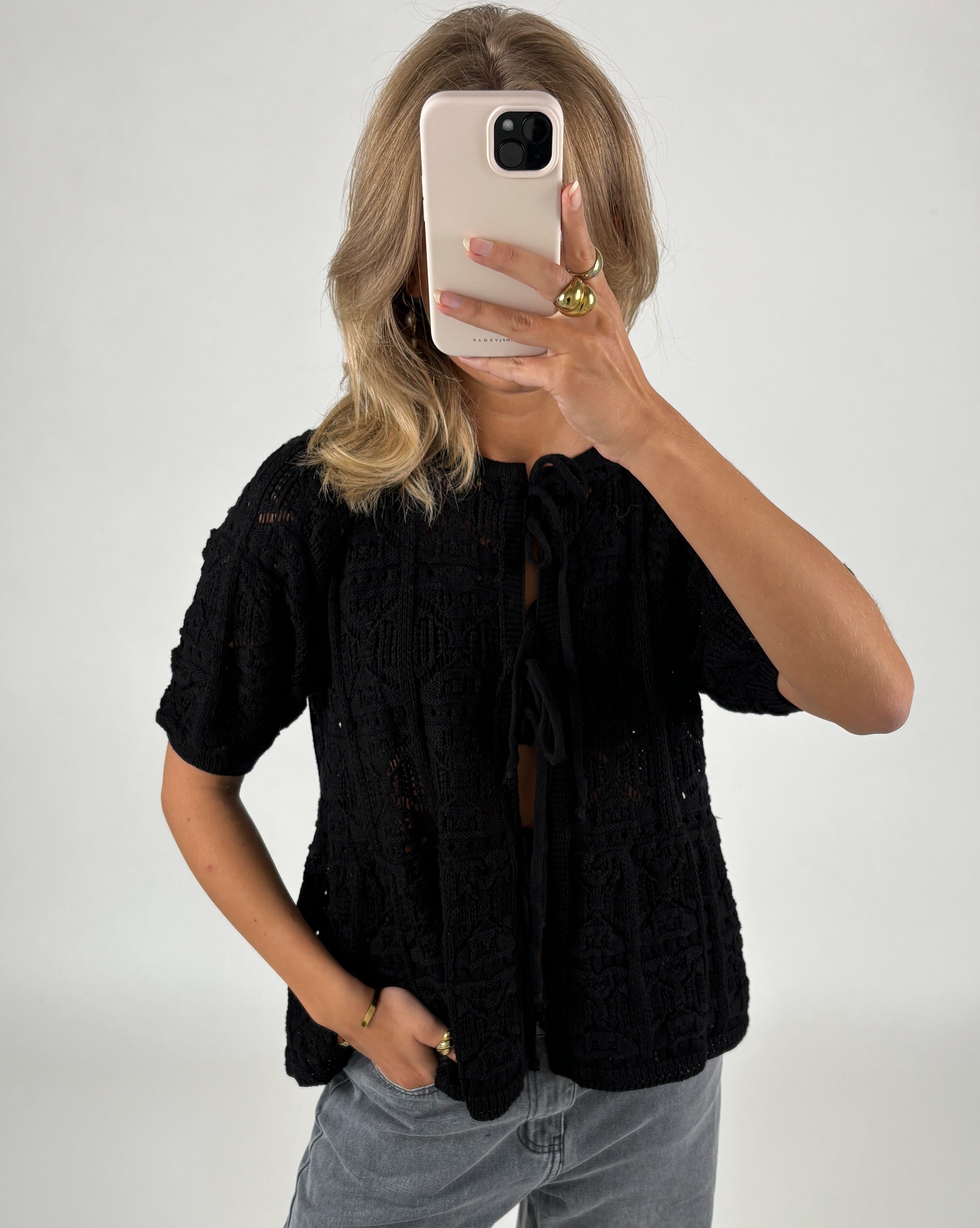 Person tager et spejlbillede iført sort striktrøje og grå jeans, med en smartphone foran ansigtet. Baggrunden er ensfarvet lys.