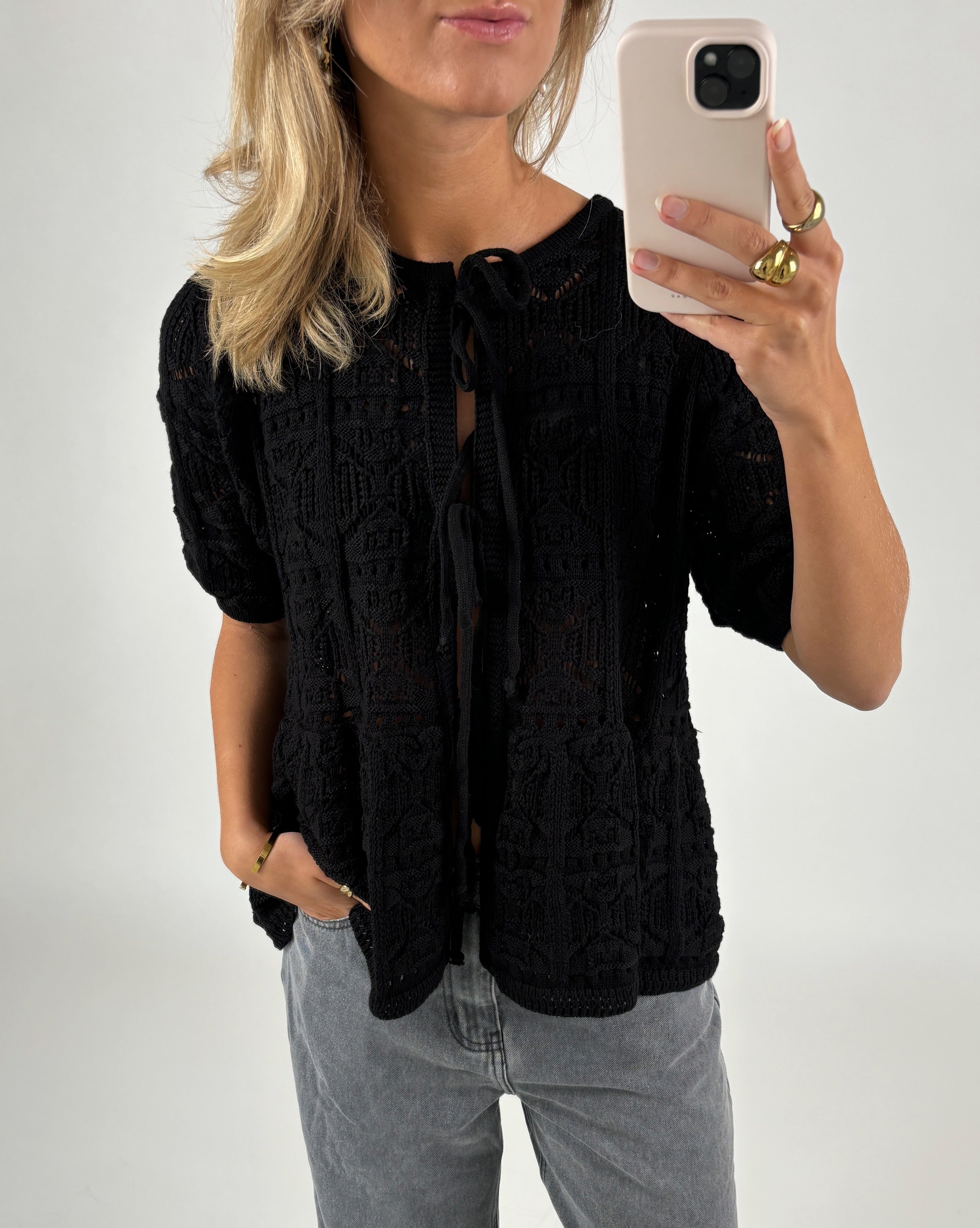 Kvinde iført sort broderet cardigan holder en smartphone, står med en hånd i lommen i et neutralt oplyst rum. Hun bærer ringsmykker på fingrene.