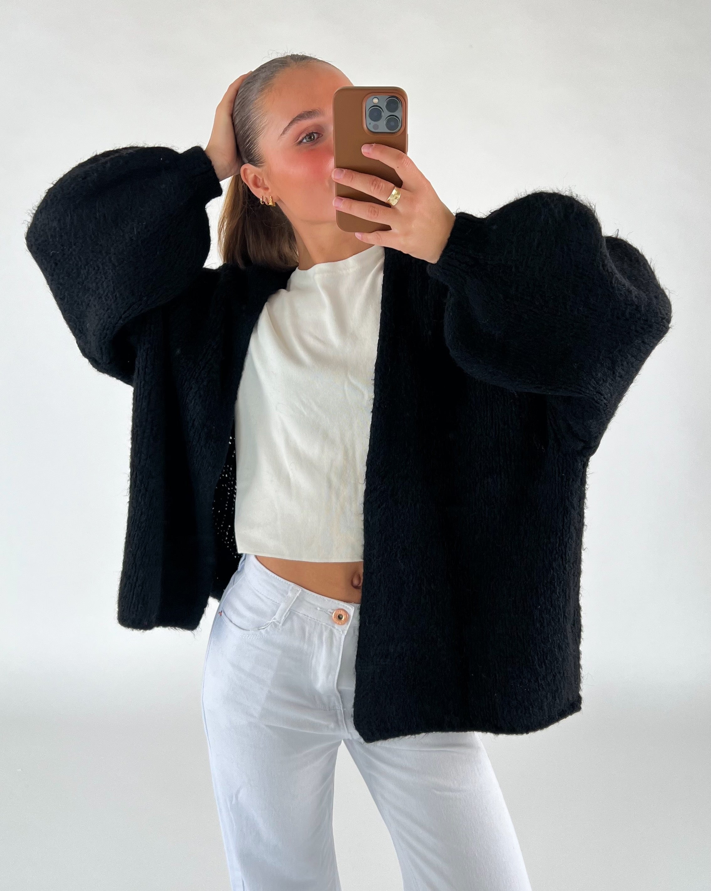 En person i sort sweater og hvide bukser tager et spejlfoto med en telefon i et minimalistisk rum. Personen holder håret tilbage med en hånd.