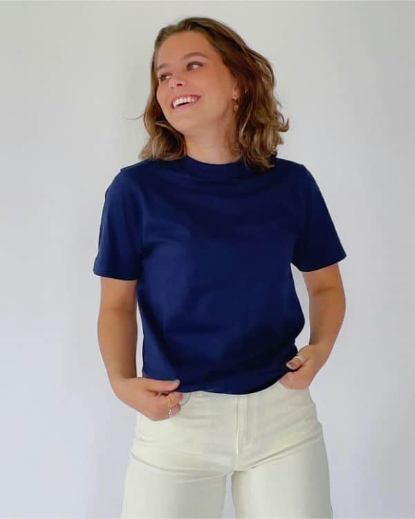En person i en mørkeblå T-shirt smiler og kigger til siden, mens de står i lyse bukser mod en hvid baggrund.