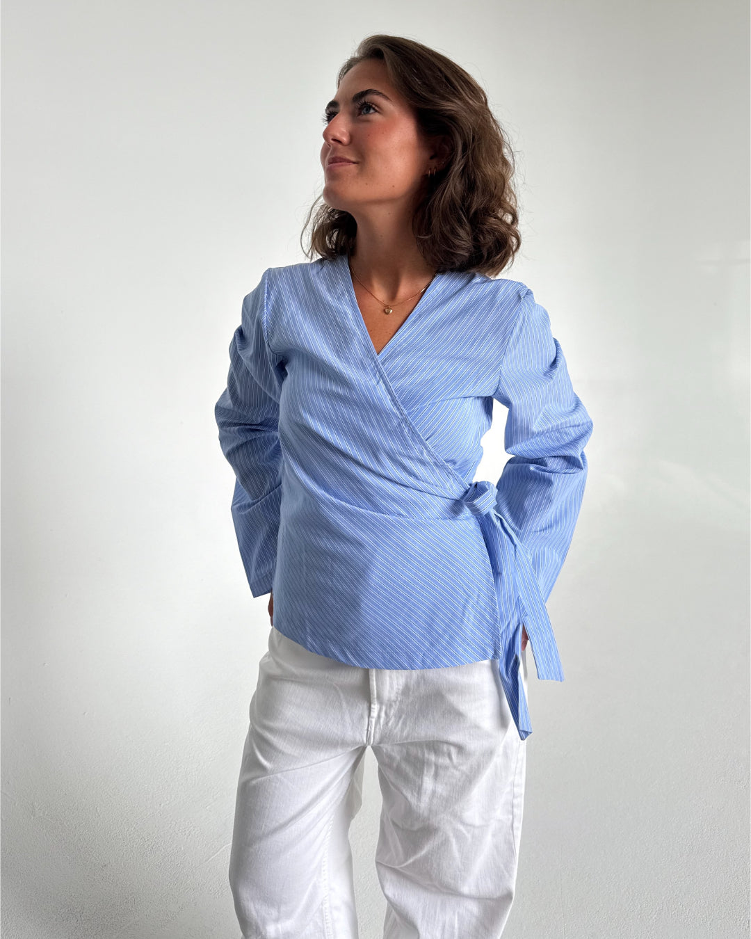 Modelfoto af pige som bærer en slå-om skjorte i lyseblå med striber, samt et par hvide Nanna jeans. Baggrund hvid. Alt fra By Sofie Sønderby
