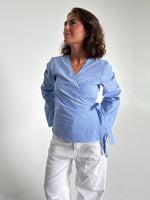 Modelfoto af pige som bærer en slå-om skjorte i lyseblå med striber, samt et par hvide Nanna jeans. Baggrund hvid. Alt fra By Sofie Sønderby