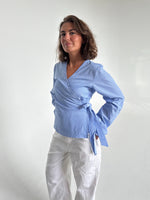 Modelfoto af pige som bærer en slå-om skjorte i lyseblå med striber, samt et par hvide Nanna jeans. Baggrund hvid. Alt fra By Sofie Sønderby