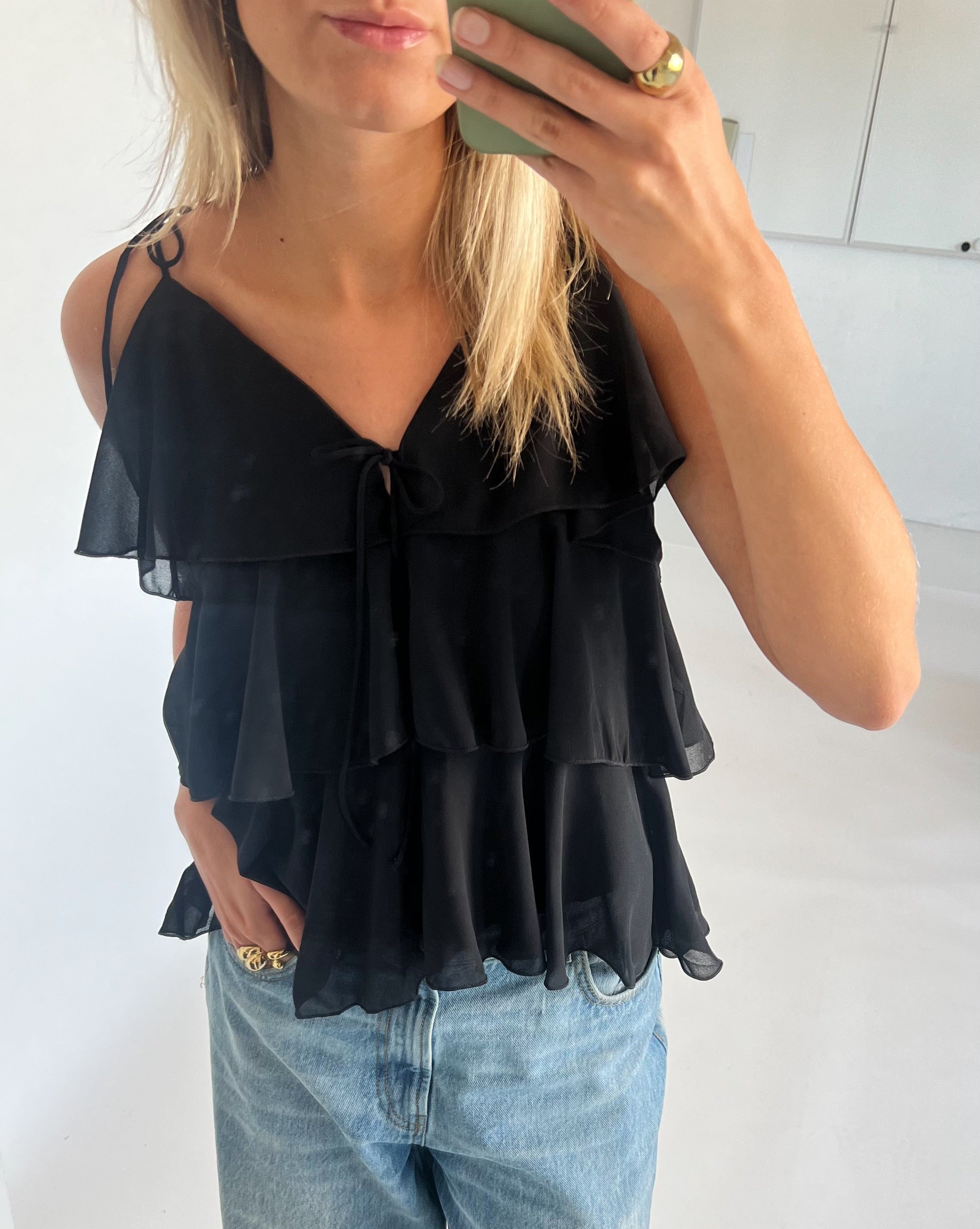 En person iført en sort, flæsebluse tager et spejlbillede, mens de holder en grøn telefon. De har også lyseblå jeans på. Rummet er lyst med hvide vægge.