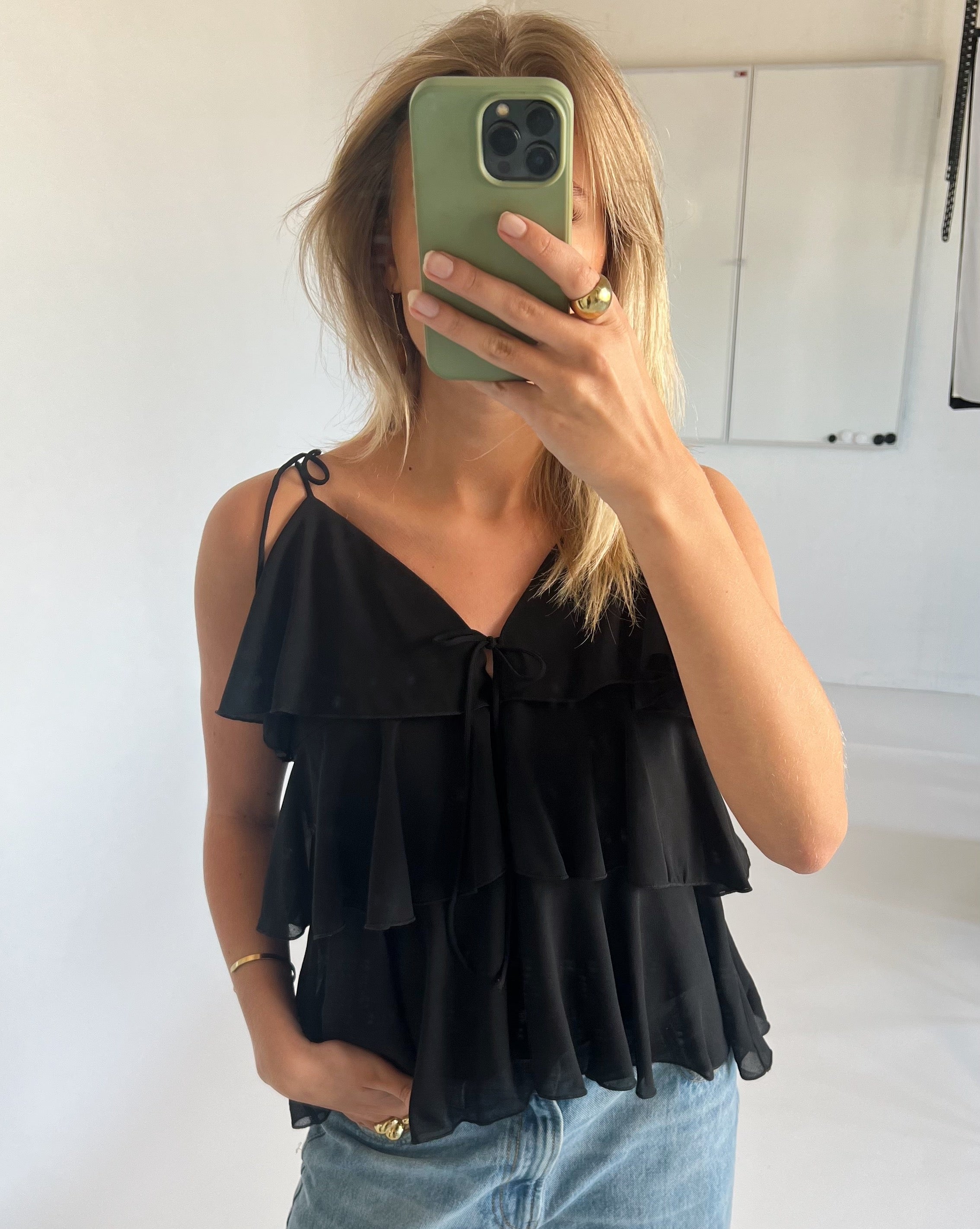 Kvinde står med en grøn telefon, dækker ansigtet, iført sort flæsetop og blå jeans, i et lyst rum med en hvid tavle i baggrunden.