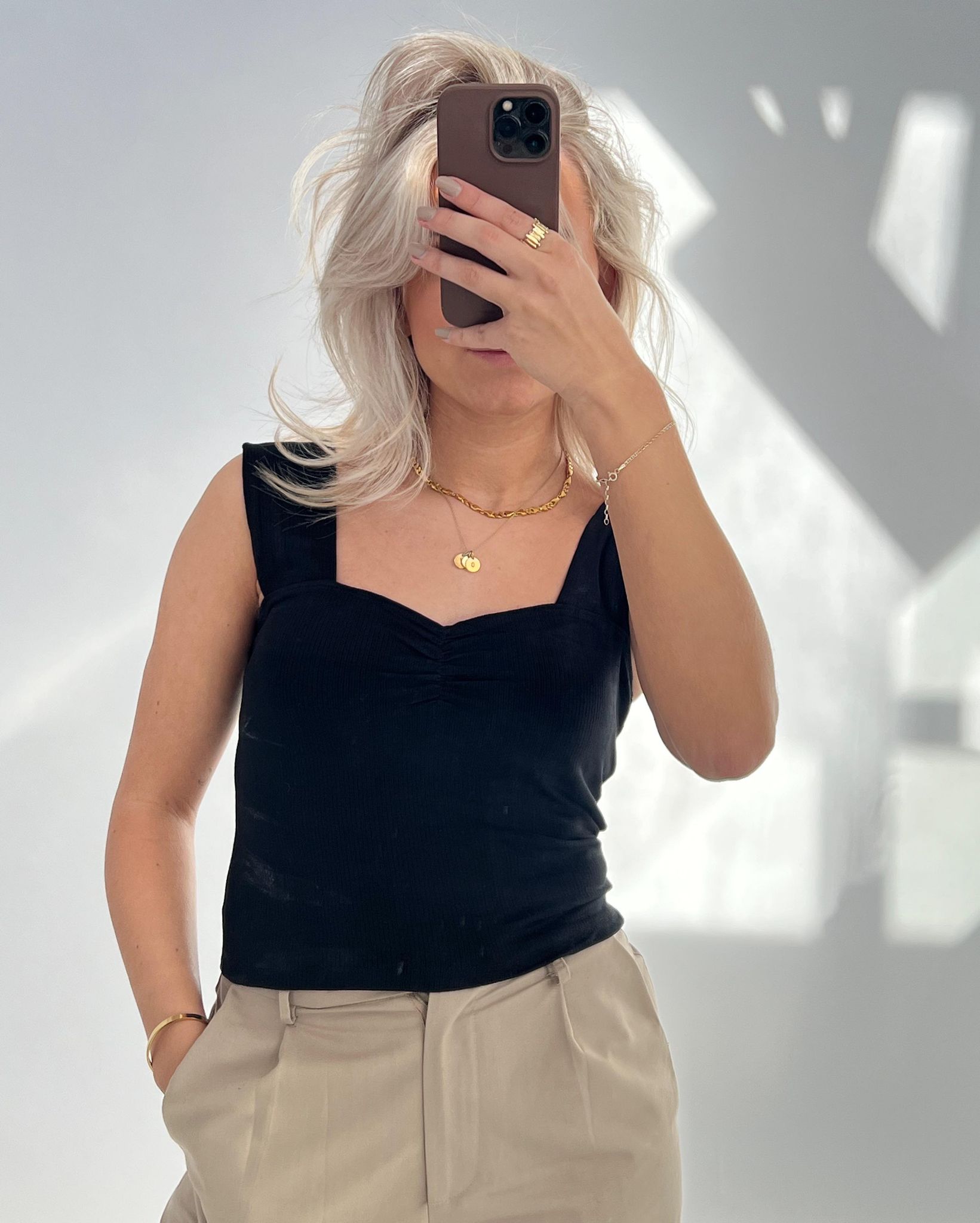 En person holder en smartphone op foran ansigtet i et rum med naturligt lys. Personen har blondt hår, sort top og beige bukser, og bærer guldsmykker.