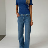 En person står iført en blå kortærmet bluse og blå jeans i et minimalistisk studie med en neutral baggrund.