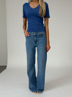 En person står iført en blå kortærmet bluse og blå jeans i et minimalistisk studie med en neutral baggrund.