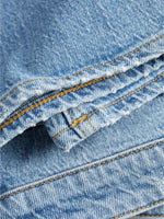 Blå denimstof ligger foldet med synlige gulbrune syninger. Stoffet er ru med let slidte kanter, hvilket er typisk for jeansmateriale.