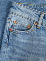 Et par blå jeans ligger fladt, med fokus på lommen dekoreret med kobbernitter. Synlige sømme og detaljer viser stoffets struktur i et enkel og tidløst design.