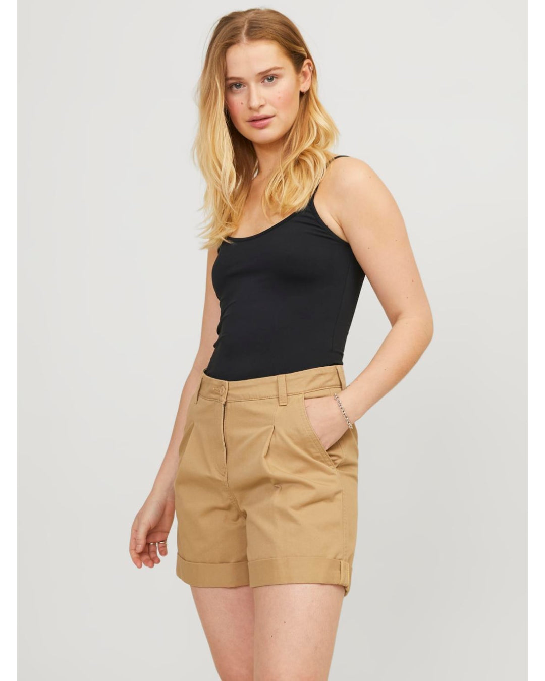 En kvinde med langt blond hår står roligt, iført sort tanktop og beige shorts. Hun hviler en hånd i lommen, med en neutral baggrund.