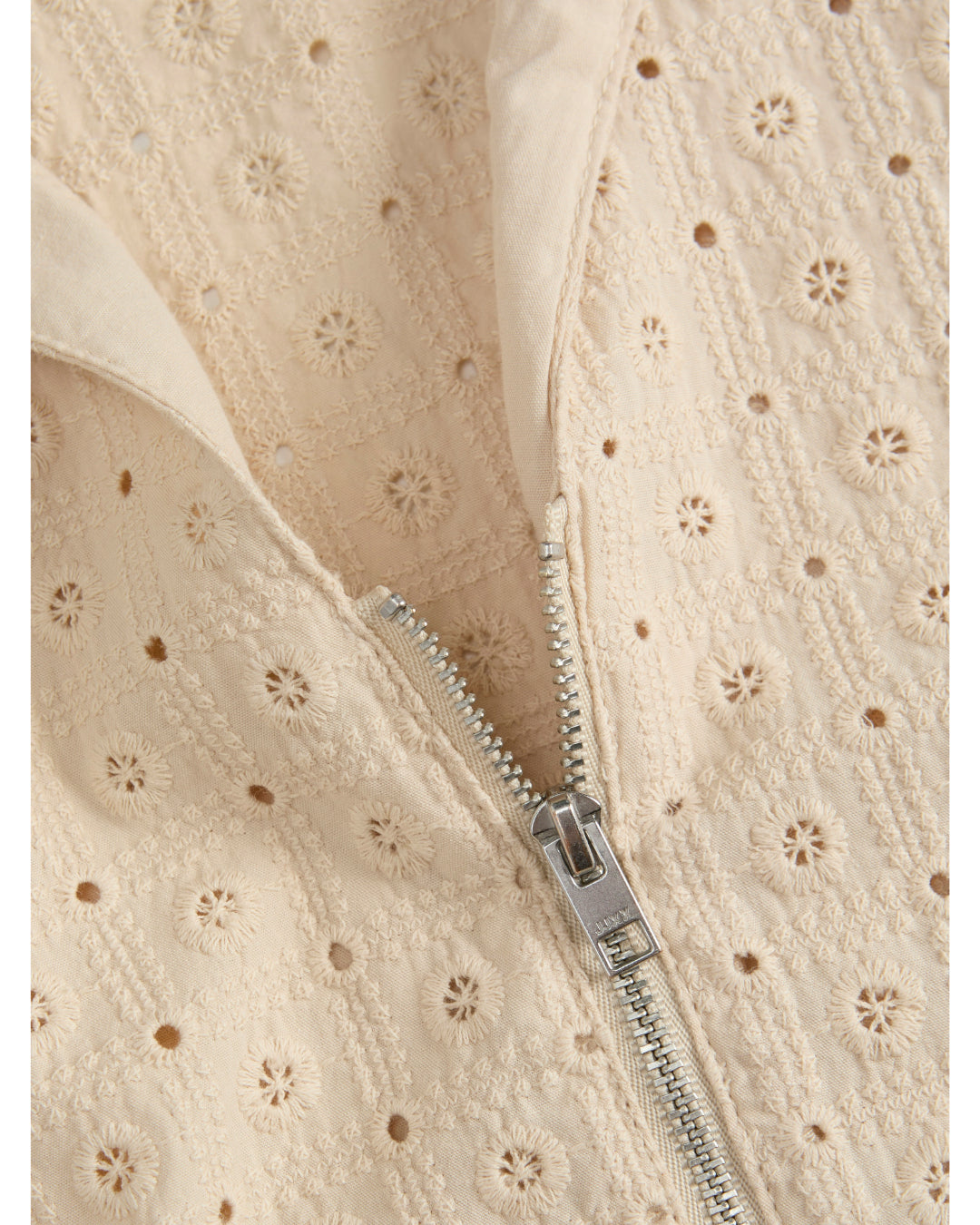 En beige stof med blomstermønster åbner sig ved hjælp af en metallynlås. Mønsteret består af små cirkulære blomster, omgivet af firkanter med broderede detaljer.
