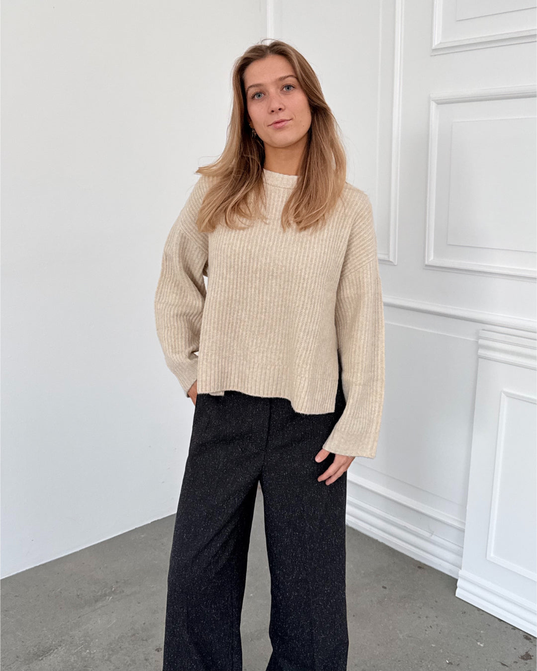 Modelfoto af pige i creme SIGGA strik, samt sortmelerede ELLIS bukser. Alt er fra BySofieSønderby.