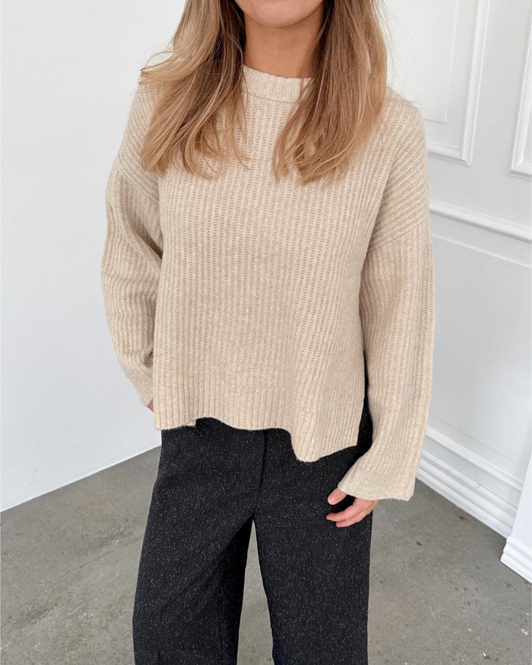 Modelfoto af pige i creme SIGGA strik, samt sortmelerede ELLIS bukser. Alt er fra BySofieSønderby.