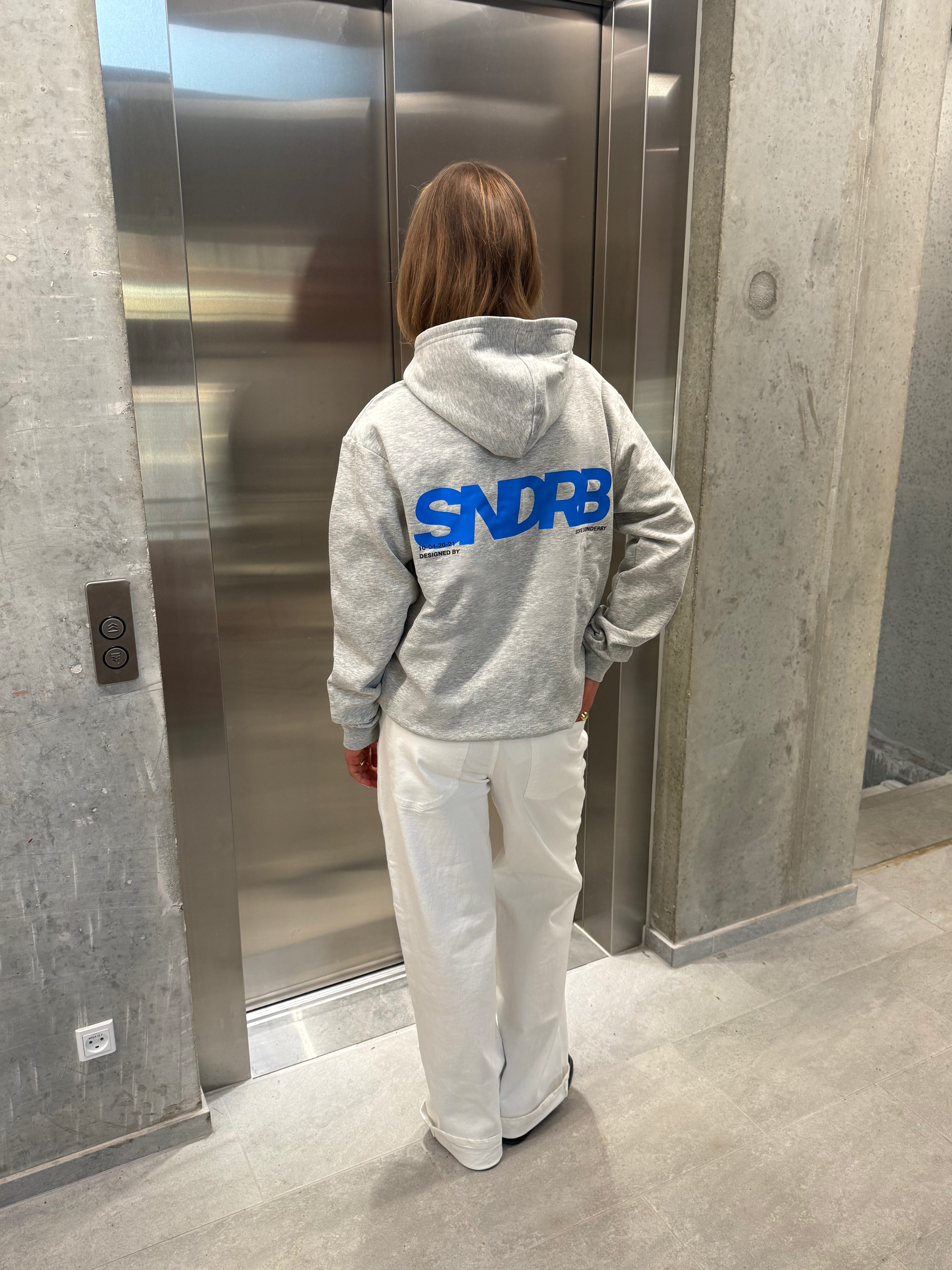 Stemningsbillede af pige der bærer en grå hoodie med blåt SNDRB tryk og hvide NANNA jeans.  Fra BySofieSønderby